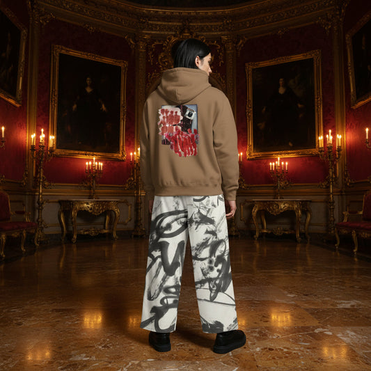A person stands in a grand, ornate room with red walls, gold-framed portraits, and candelabras, wearing a tan hoodie with a large red and black graphic on the back and white wide-leg pants with black abstract brushstroke patterns.