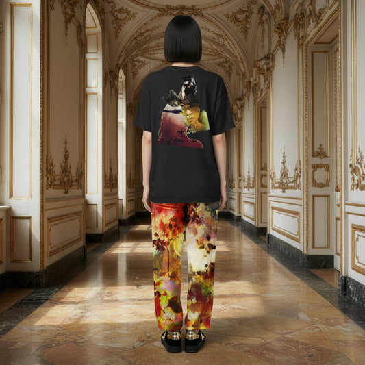 A model stands in a grand, ornate hallway with beige walls and gold detailing, viewed from behind. The model wears a black graphic t-shirt featuring a colorful print of a woman and a dog, paired with colorful abstract-patterned pants in red, yellow, green, and white.